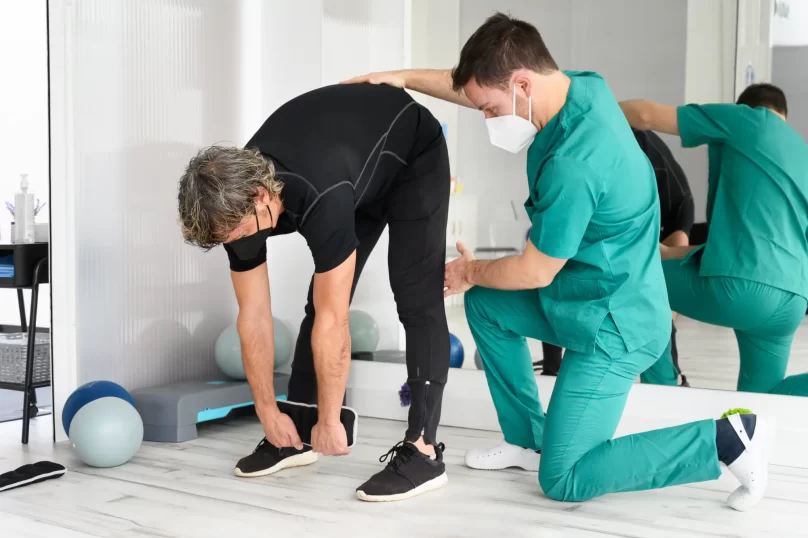 Fisioterapeuta asistiendo a un paciente en ejercicios de estiramiento en un ambiente de rehabilitación, con pelotas de ejercicio en el fondo.
