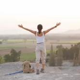 Mujer de espaldas con los brazos extendidos, disfrutando de un paisaje natural durante el amanecer, con una mochila y bastones de senderismo a su lado.