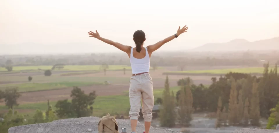 Mujer de espaldas con los brazos extendidos, disfrutando de un paisaje natural durante el amanecer, con una mochila y bastones de senderismo a su lado.
