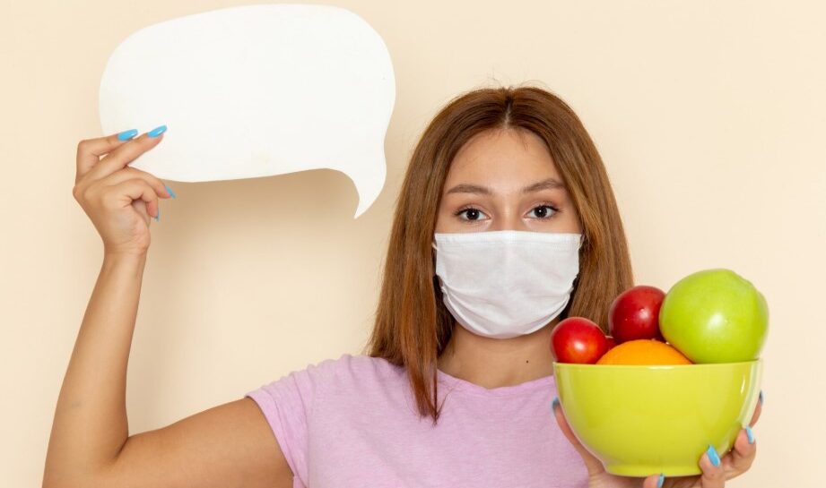 Mujer con mascarilla sosteniendo un tazón verde lleno de frutas variadas y un cartel en blanco en la otra mano, sobre fondo beige.