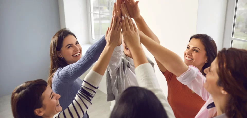 Grupo de mujeres sonriendo y levantando las manos en un gesto de celebración, en un ambiente luminoso y moderno. La imagen refleja unión y camaradería entre amigas.