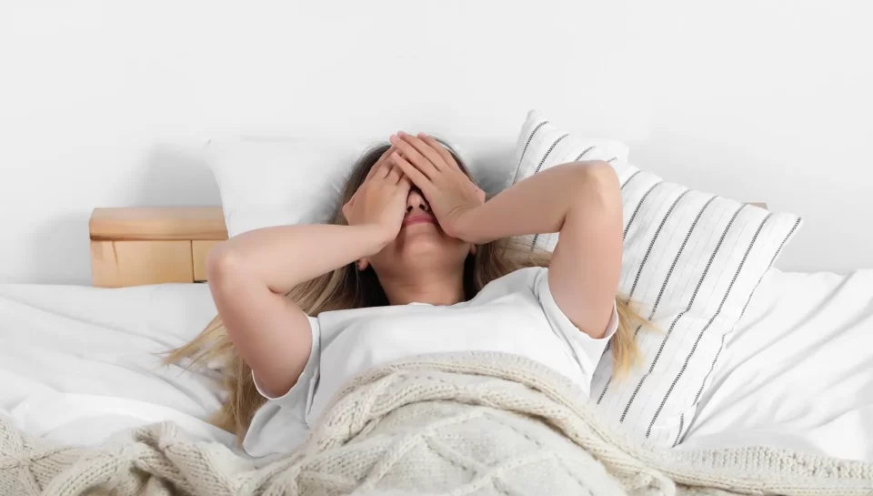 Mujer en la cama con las manos en la cara, mostrando signos de estrés o fatiga. Sábanas blancas y un cobertor de lana en un entorno relajante.